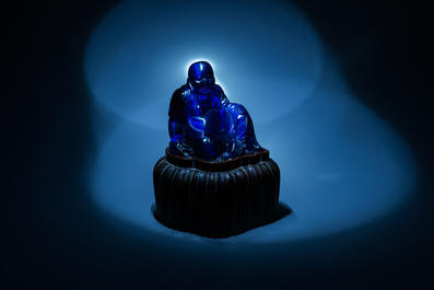 A Chinese blue Beijing glass figure of the laughing Buddha seated on a wooden stand, 19th C.